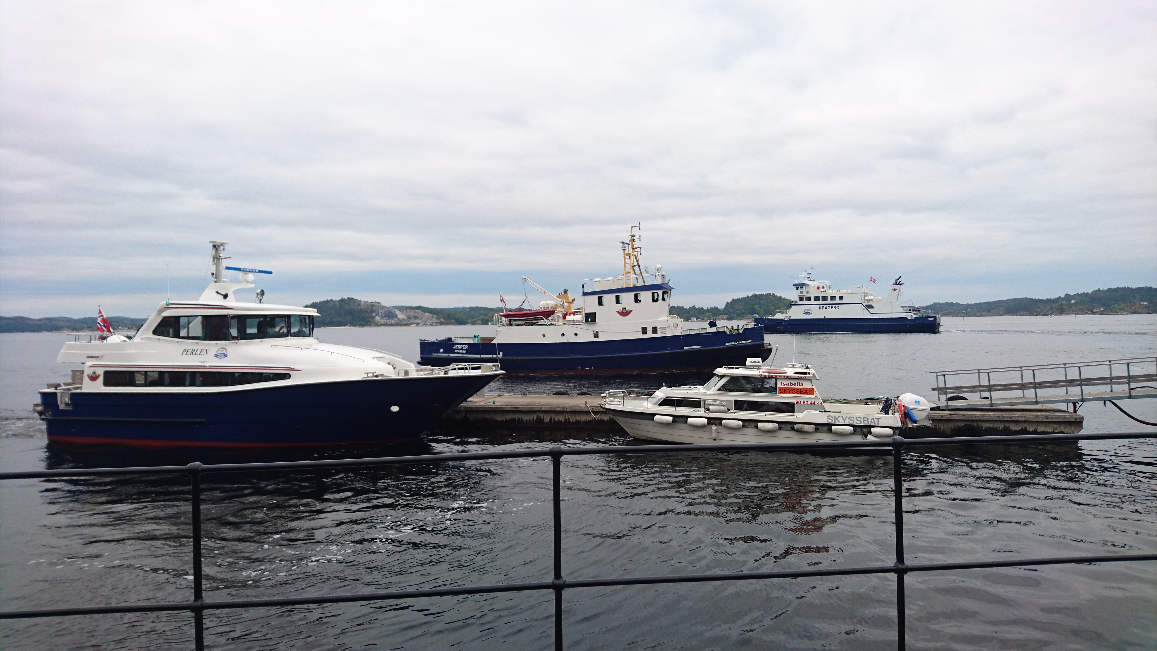 A modern office space featuring wooden desks with computers, light-colored rolling chairs, and large windows allowing natural light. Green plants are placed throughout the room, adding a fresh touch to the contemporary design.